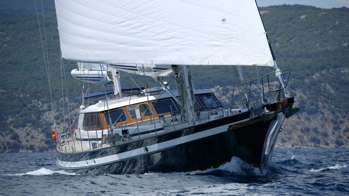 42m Jongert Infatuation sail custom yacht by Tony Castro Yachts with dark navy hull and twin masts, anchored in crystal clear turquoise waters near rocky coastline