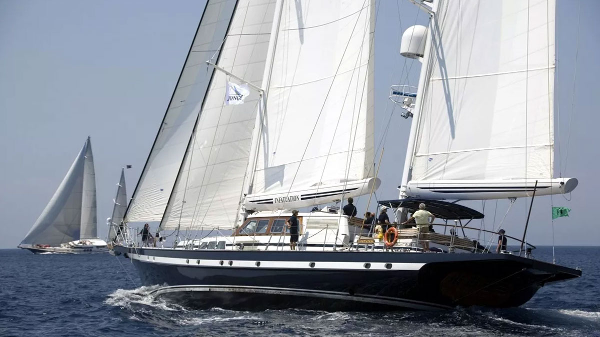 42m Jongert Infatuation sail custom yacht by Tony Castro Yachts with dark navy hull and twin masts, anchored in crystal clear turquoise waters near rocky coastline