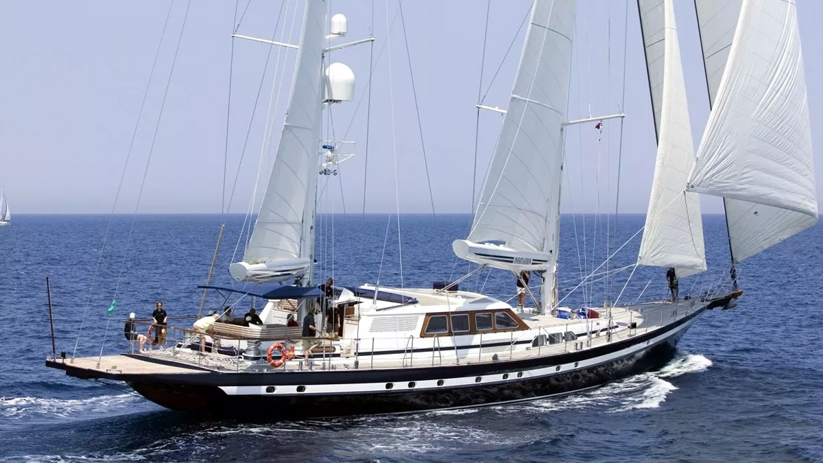 42m Jongert Infatuation sail custom yacht by Tony Castro Yachts with dark navy hull and twin masts, anchored in crystal clear turquoise waters near rocky coastline