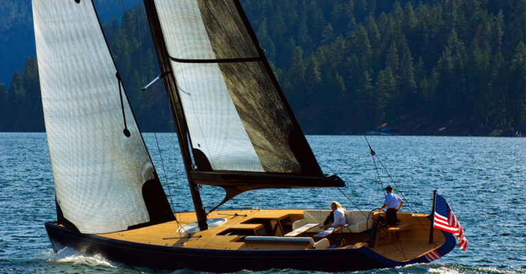 18m Sizzler custom sail yacht by Tony Castro Yachts with deep blue hull, carbon rig, and warm teak deck, sailing on a calm lake with forested hills in the background